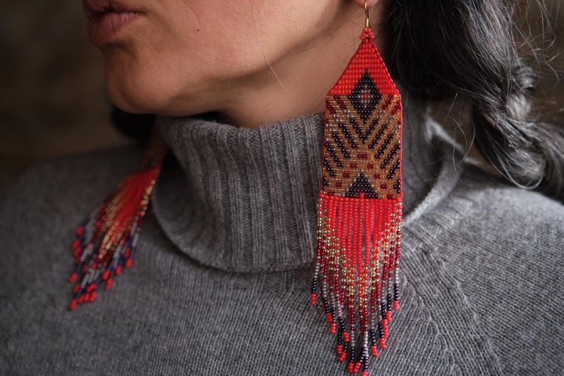 May include: Long, beaded earrings with a geometric design. The earrings feature a red, brown, and black pattern with dangling fringe. The earrings are worn with a gray turtleneck sweater.