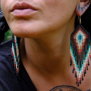 May include: A pair of long, beaded earrings with a geometric pattern in shades of black, red, green, blue, and white. The earrings are hanging from the ears of a person with long black hair.