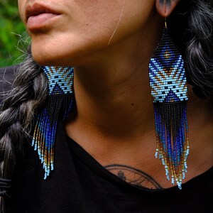 May include: Handmade beaded earrings with a geometric design. The earrings feature a pattern of light blue, dark blue, and gold beads, with black fringe. The earrings are long and dangle, adding a touch of elegance.