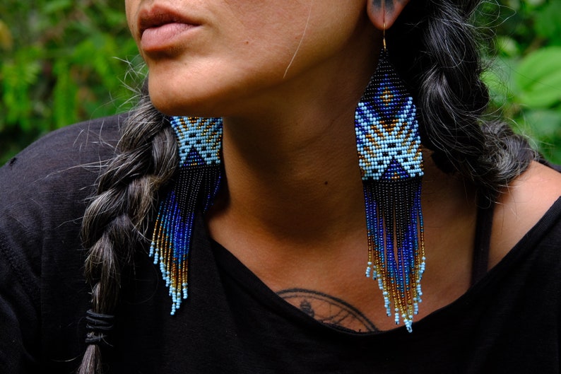 May include: Handmade beaded earrings with a geometric design. The earrings feature a pattern of light blue, dark blue, and gold beads, with black fringe. The earrings are long and dangle, adding a touch of elegance.