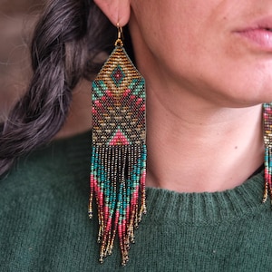 May include: Close-up of beaded earrings with a geometric design. The earrings feature a mix of colors, including teal, pink, gold, and black. The earrings have a rectangular top and long fringe. The earrings are hanging from gold-colored hooks.