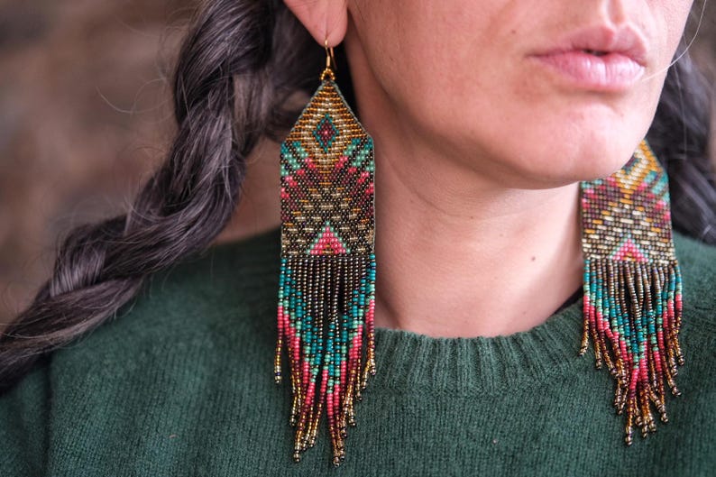 May include: Close-up of beaded earrings with a geometric design. The earrings feature a mix of colors, including teal, pink, gold, and black. The earrings have a rectangular top and long fringe. The earrings are hanging from gold-colored hooks.