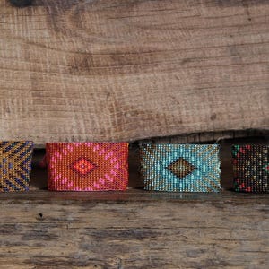 Könnte beinhalten: Vier Armbänder mit geometrischen Mustern. Die Armbänder sind in Blau-, Orange- und Brauntönen gehalten und vor einem Holzgrund platziert. Jedes Armband hat ein einzigartiges Design, das aufwendige Perlenarbeiten zeigt.