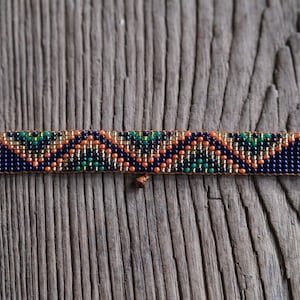 Op de afbeelding: Een kralenarmband met een geometrisch patroon in marineblauw, oranje, groen en goud. De armband is rechthoekig met afgeronde uiteinden en wordt gepresenteerd op een verweerd houten oppervlak.