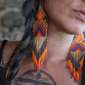 May include: Long, beaded earrings in shades of orange, yellow, purple, and gold. The earrings have a geometric design and a fringe at the bottom. The earrings are hanging from the ear.