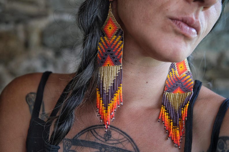 May include: Long, beaded earrings in shades of orange, yellow, purple, and gold. The earrings have a geometric design and a fringe at the bottom. The earrings are hanging from the ear.
