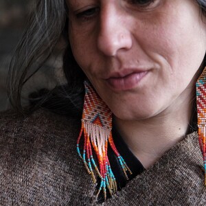 May include: Handmade beaded earrings with a geometric design. The earrings feature a pattern of orange, red, turquoise, and white beads, with a fringe of beads in similar colors. The earrings are suspended from gold-colored hooks.