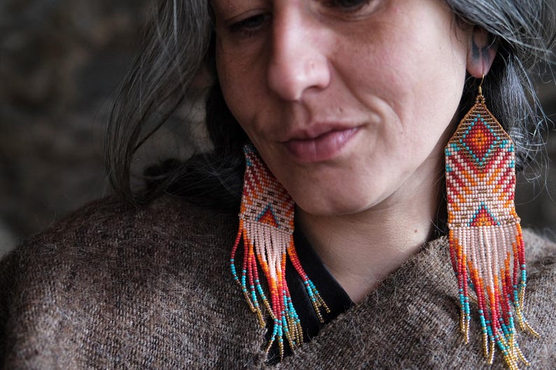 May include: Handmade beaded earrings with a geometric design. The earrings feature a pattern of orange, red, turquoise, and white beads, with a fringe of beads in similar colors. The earrings are suspended from gold-colored hooks.