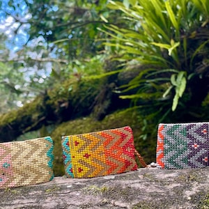 May include: Three beaded clutch purses in pink, yellow-orange, and green-purple color schemes. The purses are rectangular with intricate geometric patterns. They are displayed on a mossy log with a lush green background.
