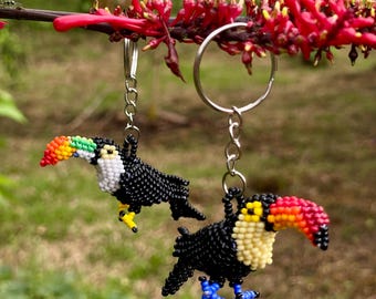 May include: Two beaded toucan keychains. The toucans have black bodies, white chests, and colorful beaks in red, orange, yellow, and green. The feet are yellow and blue. The keychains hang from a silver ring and chain.