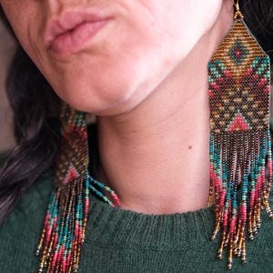 May include: Handmade beaded earrings with a geometric design. The earrings feature a combination of gold, teal, pink, and brown beads. The earrings have a rectangular top with a fringe of beads hanging down. The earrings are worn by a person wearing a green sweater.