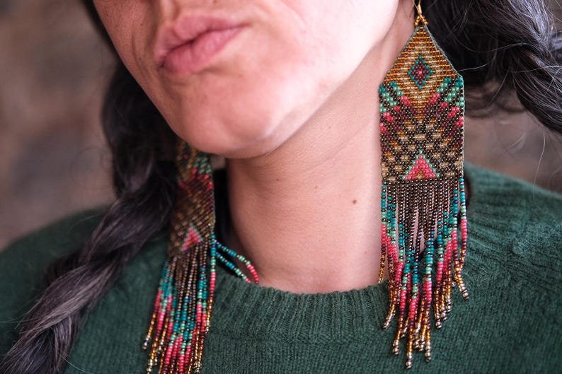 May include: Handmade beaded earrings with a geometric design. The earrings feature a combination of gold, teal, pink, and brown beads. The earrings have a rectangular top with a fringe of beads hanging down. The earrings are worn by a person wearing a green sweater.