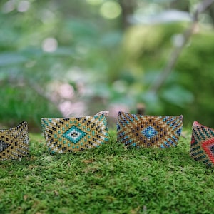 Pode incluir: Quatro pulseiras de contas com padrões geométricos em várias cores, incluindo dourado, verde-azulado, castanho e vermelho. As pulseiras são exibidas sobre uma cama de musgo verde, com um fundo natural desfocado.