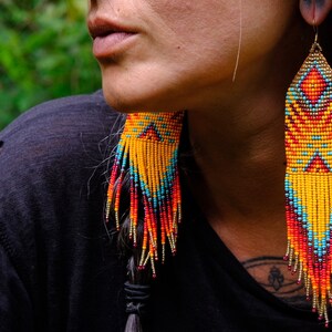 May include: Long, beaded earrings in shades of yellow, orange, red, and turquoise. The earrings have a geometric pattern and fringe. The earrings are hanging from gold-colored hooks.