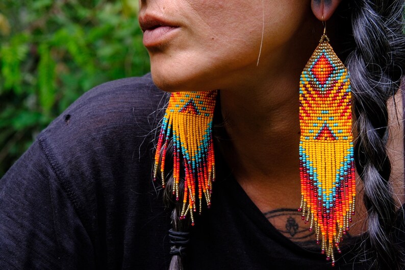 May include: Long, beaded earrings in shades of yellow, orange, red, and turquoise. The earrings have a geometric pattern and fringe. The earrings are hanging from gold-colored hooks.
