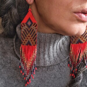 May include: Red, brown, and gold beaded earrings with a geometric pattern and fringe. The earrings have a triangular top and a rectangular body with a detailed design. The fringe is made of red, gold, and silver beads.