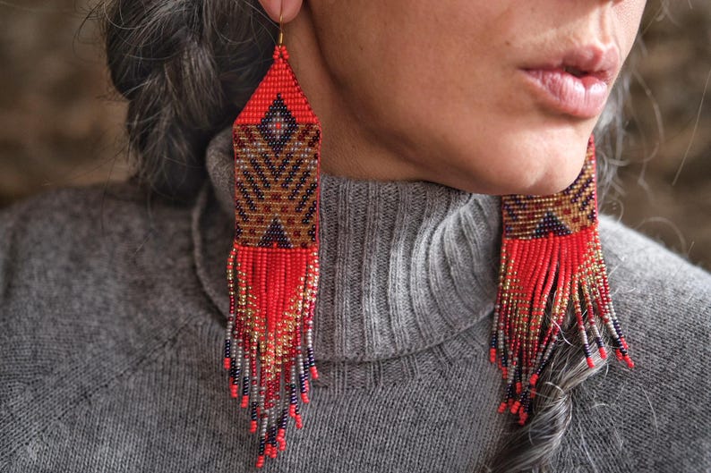 May include: Red, brown, and gold beaded earrings with a geometric pattern and fringe. The earrings have a triangular top and a rectangular body with a detailed design. The fringe is made of red, gold, and silver beads.