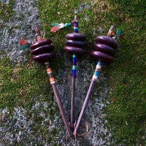 Op de afbeelding: Drie spintollen met donkerbruine houten stelen en kransen. Elke spintol heeft een stapel van drie donkerbruine ronde gewichten, kleurrijke draadomwikkelingen op de stelen en kleine decoratieve veren bevestigd aan de bovenste uiteinden.