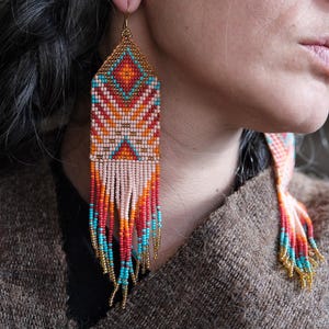 May include: Handmade beaded earrings with a geometric design. The earrings feature a diamond-shaped top with a pattern of orange, turquoise, and white beads. The bottom of the earrings have long fringe in shades of red, orange, and turquoise.