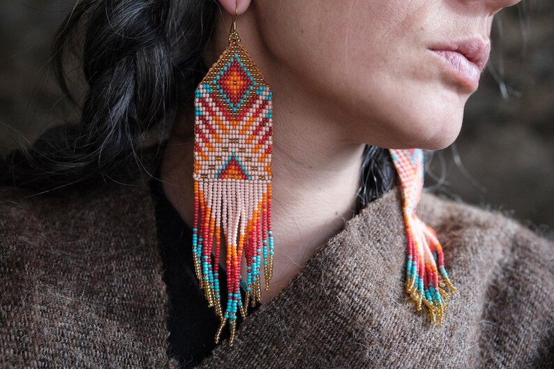 May include: Handmade beaded earrings with a geometric design. The earrings feature a diamond-shaped top with a pattern of orange, turquoise, and white beads. The bottom of the earrings have long fringe in shades of red, orange, and turquoise.