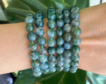 May include: A stack of green agate beaded bracelets with a mottled pattern. The bracelets are worn on a person's wrist.