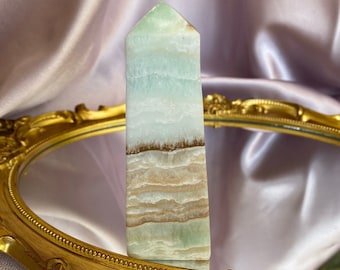 May include: A green and white banded crystal point, also known as a crystal obelisk, sits on a gold-colored tray with a mirrored surface.