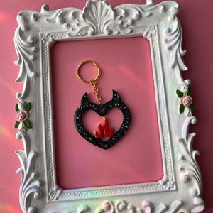 May include: Black heart-shaped keychain with a red flame design inside. The keychain has a gold ring and is displayed in a white ornate frame.