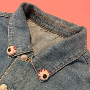 May include: Light blue denim jacket with two eyeball-shaped buttons on the collar. The buttons are white with red veins and a black pupil. The jacket has silver buttons and a classic collar.