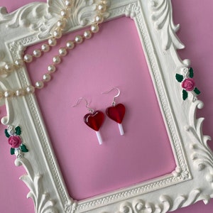 May include: A pair of heart-shaped red lollipop earrings with white sticks, displayed on a pink background within a white ornate frame. A pearl necklace is draped across the frame. The earrings are a fun, playful accessory.