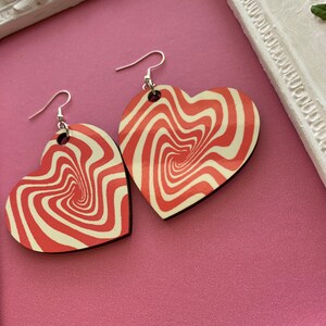 Peut inclure: Une paire de boucles d'oreilles en forme de c&oelig;ur avec un motif rouge et blanc tourbillonnant. Les boucles d'oreilles sont en bois et ont des crochets argent&eacute;s.
