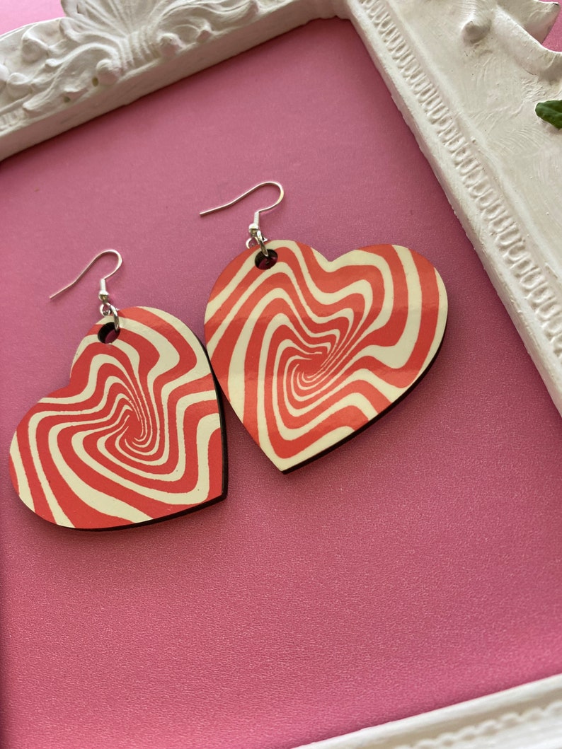 Peut inclure: Une paire de boucles d'oreilles en forme de c&oelig;ur avec un motif rouge et blanc tourbillonnant. Les boucles d'oreilles sont en bois et ont des crochets argent&eacute;s.