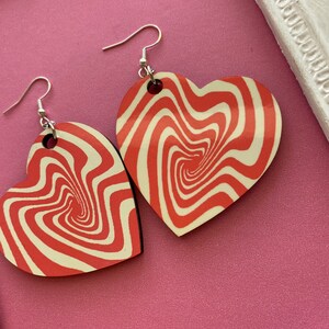 Peut inclure: Une paire de boucles d'oreilles en forme de c&oelig;ur rouge et blanc avec un motif tourbillonnant. Les boucles d'oreilles sont en bois et ont des crochets argent&eacute;s.
