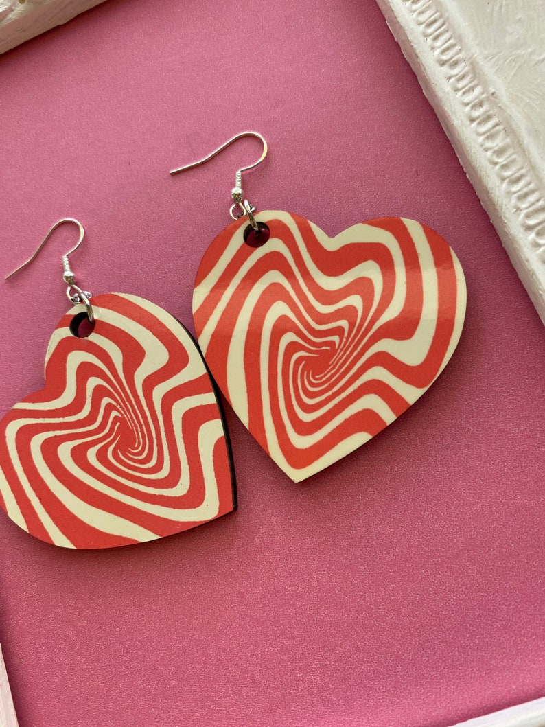 Peut inclure: Une paire de boucles d'oreilles en forme de c&oelig;ur rouge et blanc avec un motif tourbillonnant. Les boucles d'oreilles sont en bois et ont des crochets argent&eacute;s.