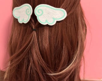 May include: Two white felt hair clips in the shape of angel wings, each outlined in light green. The hair clips are attached to brown hair, styled in a half-up, half-down hairstyle. The background is a solid pink color.