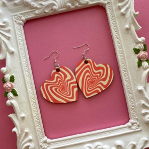 Peut inclure: Une paire de boucles d'oreilles en forme de c&oelig;ur rouge et blanc avec un motif tourbillonnant. Les boucles d'oreilles sont suspendues &agrave; des crochets argent&eacute;s.