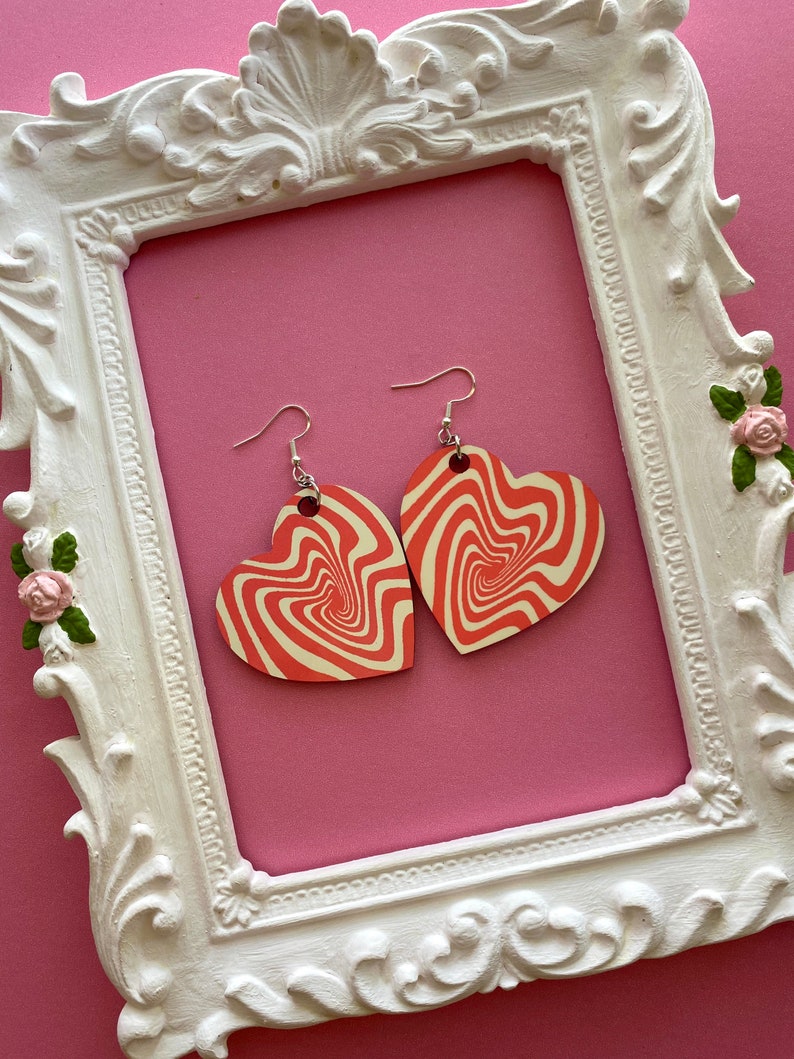 Peut inclure: Une paire de boucles d'oreilles en forme de c&oelig;ur rouge et blanc avec un motif tourbillonnant. Les boucles d'oreilles sont suspendues &agrave; des crochets argent&eacute;s.