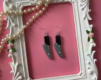 Peut inclure: Une paire de boucles d'oreilles en forme de couteau avec des paillettes noires et argentées. Les boucles d'oreilles sont suspendues à un cadre photo blanc sur lequel est drapé un collier de perles.