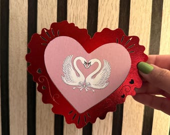 Peut inclure: Une carte rouge en forme de cœur avec un cœur intérieur rose représentant deux cygnes formant un cœur. La carte a un bord festonné et des découpes décoratives. Les cygnes sont blancs avec des ailes détaillées.