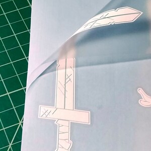 Pu&ograve; includere: Un foglio di adesivi in ​​vinile bianco con un design a forma di spada, piuma e mano. Il foglio &egrave; su un tappetino da taglio verde.