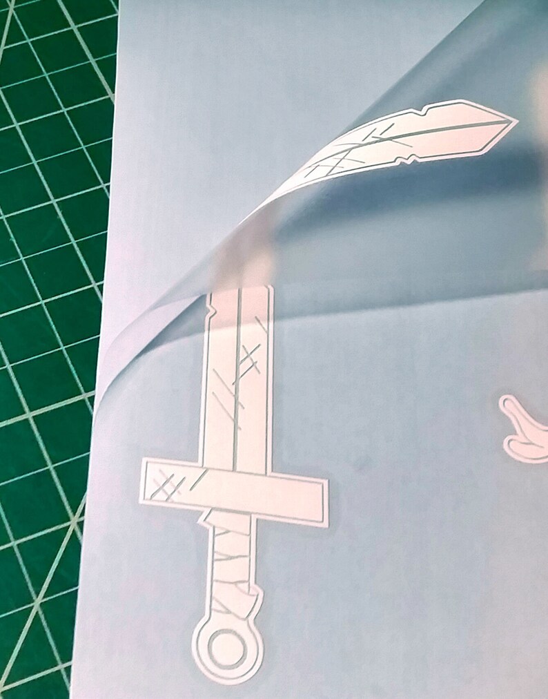 Pu&ograve; includere: Un foglio di adesivi in ​​vinile bianco con un design a forma di spada, piuma e mano. Il foglio &egrave; su un tappetino da taglio verde.