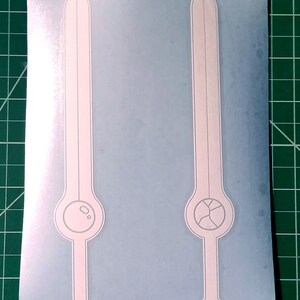Pu&ograve; includere: Due adesivi in ​​vinile bianco di una spada con un design circolare alla base. La spada a sinistra ha un cerchio pieno, mentre la spada a destra ha un cerchio con una crepa.
