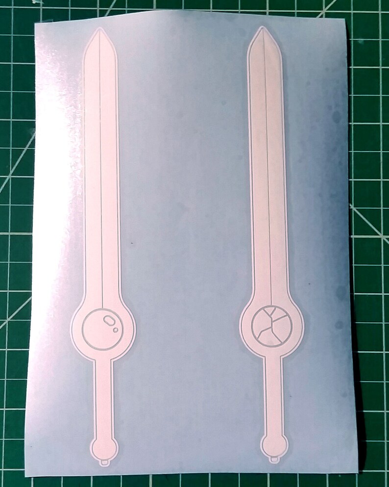 Pu&ograve; includere: Due adesivi in ​​vinile bianco di una spada con un design circolare alla base. La spada a sinistra ha un cerchio pieno, mentre la spada a destra ha un cerchio con una crepa.