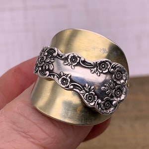 May include: A silver spoon ring with a floral design, set in a brass band. The ring is worn on a finger.