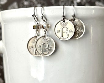 May include: Two pairs of silver-toned coin earrings featuring the letter B, a crown emblem, and the years 1964 and 1972. One pair includes small clear faceted beads on the ear wires, while the other pair consists of simple silver-toned metal hooks.