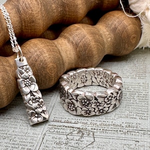 Puede incluir: Un collar y un anillo de plata con un diseño floral. El collar presenta un colgante con un patrón floral vertical, mientras que el anillo es una banda ancha con un patrón floral circular. Ambas piezas se muestran sobre una superficie de madera.