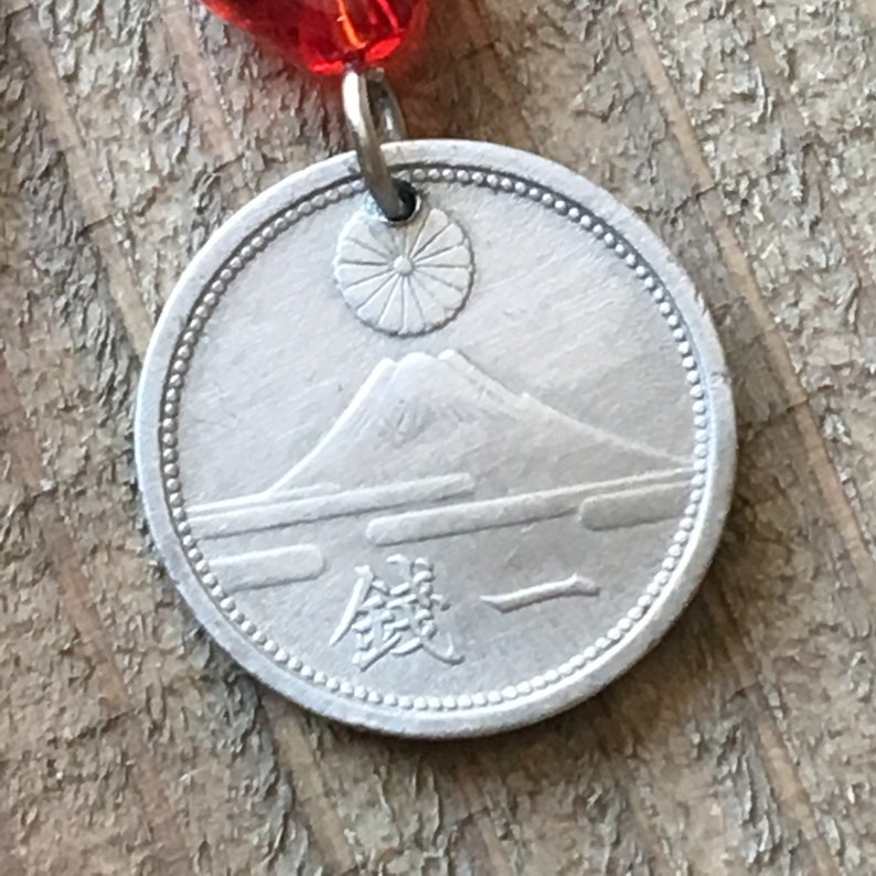 May include: A silver coin with a depiction of Mount Fuji and a rising sun. The coin has a raised rim and the Japanese word "銭" (sen) is inscribed on the bottom.