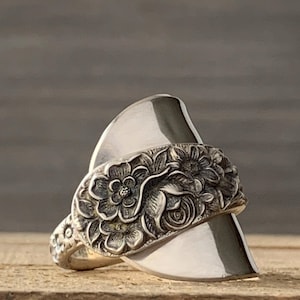 May include: A silver-toned ring crafted from a repurposed spoon, featuring a prominent bowl section that curves upward. The band displays an intricate, raised floral relief pattern with detailed petals and leaves across the front surface.