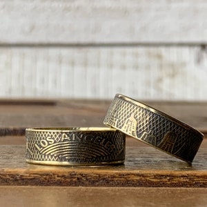 May include: Two brass-colored rings with etched designs. One ring displays the text "LOS ANGELES" above a curved line pattern. The other ring features a cityscape design. Both rings have a textured, cross-hatched pattern.