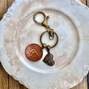 Pode incluir: Um chaveiro de bronze com uma moeda australiana de 1951 com um canguru e as palavras "Australia" e "Penny". Um amuleto em forma de coração com um design decorativo pendura no chaveiro.