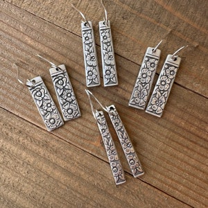 Może przedstawiać: Sześć par srebrnych kolczyków wiszących z kwiatowymi wzorami. Każdy kolczyk ma prostokątny wisiorek z detalicznym wzorem kwiatowym.